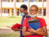 Fully Funded Scholarships for Nigerian Students in 2025 Two students wearing face masks stand outside a school building. The smiling woman in front, who dreams of fully funded scholarships for Nigerian students, holds books and a notebook, while the man behind her reads. Both appear relaxed and studious.