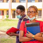 Two students wearing face masks stand outside a school building. The smiling woman in front, who dreams of fully funded scholarships for Nigerian students, holds books and a notebook, while the man behind her reads. Both appear relaxed and studious.