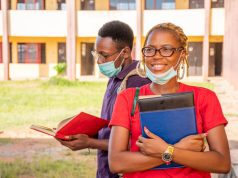 Fully Funded Scholarships for Nigerian Students in 2025 Two students wearing face masks stand outside a school building. The smiling woman in front, who dreams of fully funded scholarships for Nigerian students, holds books and a notebook, while the man behind her reads. Both appear relaxed and studious.