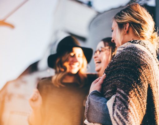 How Social Media Can Help You Find International Scholarships Three women stand close together outdoors, laughing and smiling warmly. The bright, soft lighting creates a cheerful vibe—perfect for sharing social media scholarships news with friends. One woman wears a black hat, another has a blanket around her shoulders.
