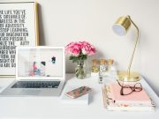 Top 5 Tips for Writing an Engaging Scholarship Blog Post A tidy desk with a laptop, pink flowers in a vase, a gold lamp, notebooks for writing an engaging scholarship blog post, glasses, a phone, and jars. A framed inspirational quote leans against the wall in the background.