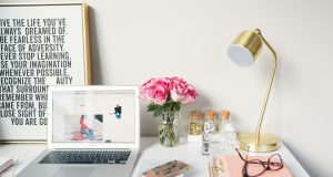 Top 5 Tips for Writing an Engaging Scholarship Blog Post A tidy desk with a laptop, pink flowers in a vase, a gold lamp, notebooks for writing an engaging scholarship blog post, glasses, a phone, and jars. A framed inspirational quote leans against the wall in the background.