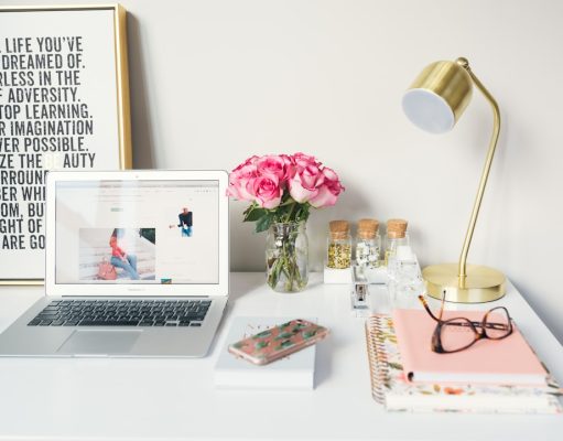 Top 5 Tips for Writing an Engaging Scholarship Blog Post A tidy desk with a laptop, pink flowers in a vase, a gold lamp, notebooks for writing an engaging scholarship blog post, glasses, a phone, and jars. A framed inspirational quote leans against the wall in the background.