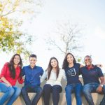 Five people sit side by side on a low wall outdoors, smiling at the camera—perhaps celebrating their success with ECE Scholarships for International Students. Trees and a clear sky are visible in the background.