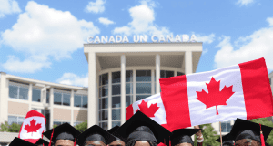 Fully Funded Scholarship in Canada for African students A diverse group of graduates in caps and gowns smile and hold Canadian flags in front of a building with Canada written on it, celebrating their achievement—thanks to a fully funded scholarship in Canada for African students—on a sunny day.